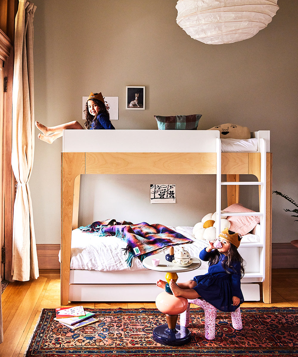 Two kids in crowns enjoy a cozy room featuring the Perch Twin Bunk Bed, toys, and a colorful rug. One child sits while the other climbs.