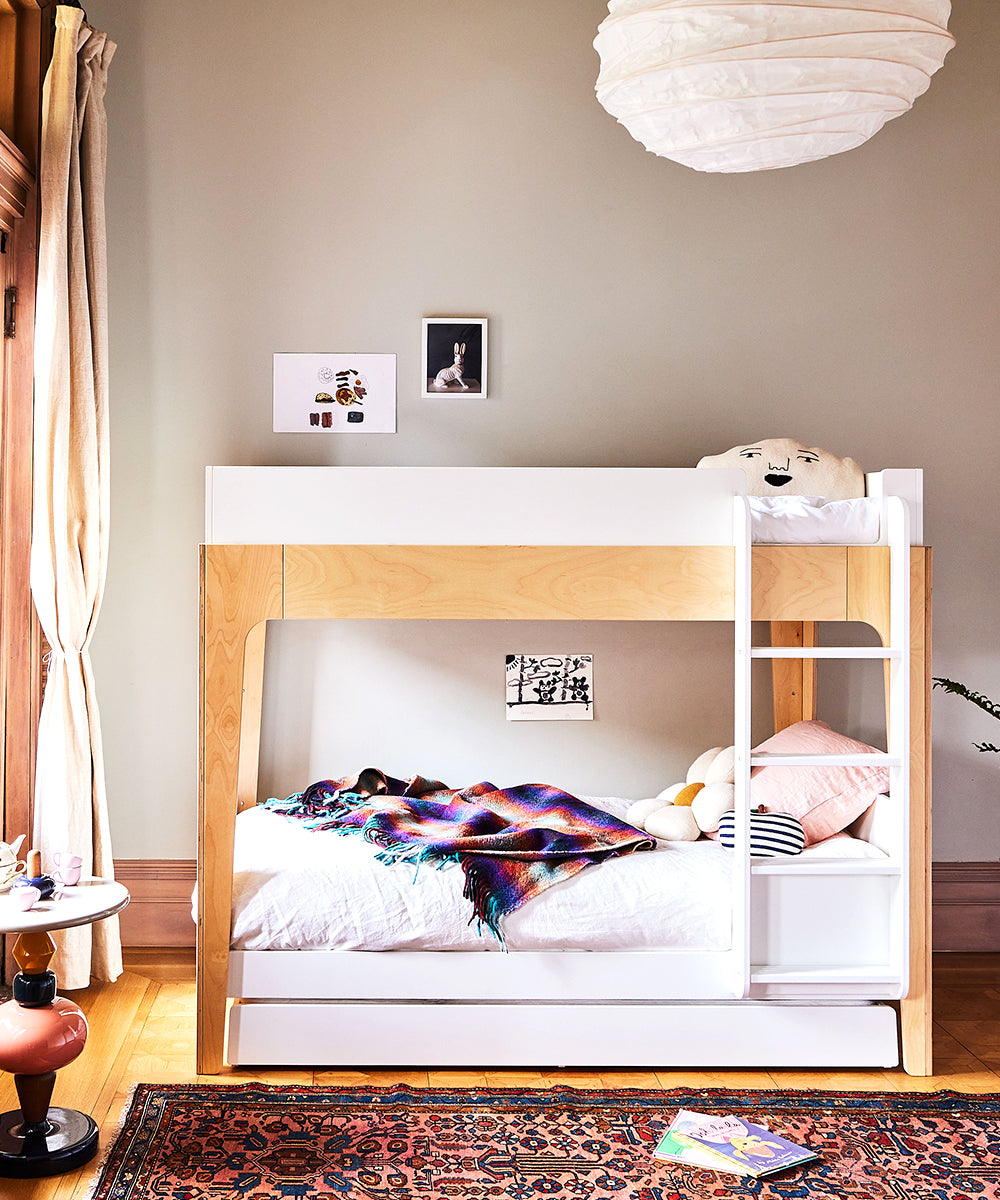 The Perch Trundle Bed & Ladder Conversion Kit serves as a childrens bunk bed, adorned with a colorful blanket and pillows, complemented by vibrant wall art. A pendant lamp illuminates the space above a patterned rug.