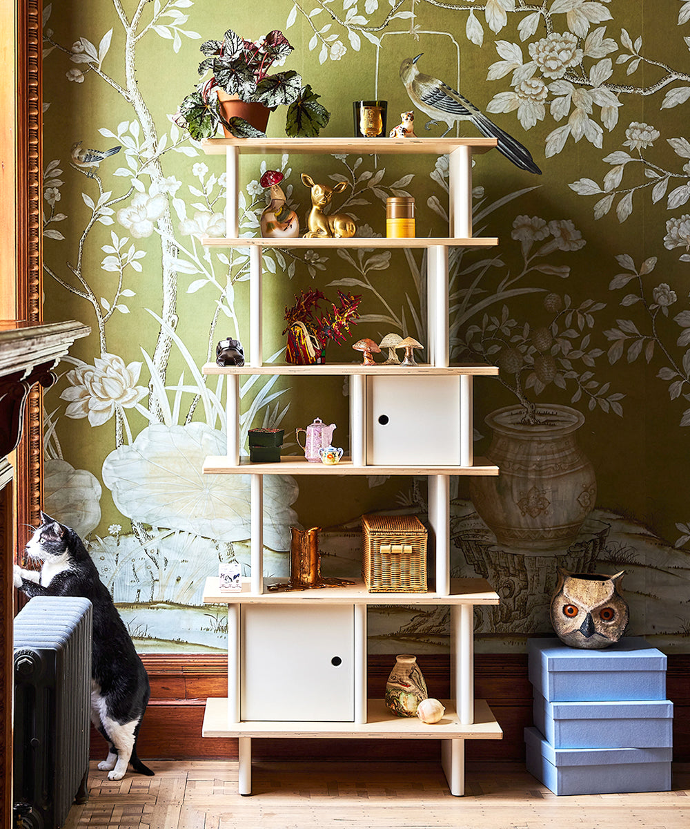 The Vertical Mini Library, styled with plants, animal figurines, and blue boxes, is set against a floral wallpaper. A cat peeks by the radiator.