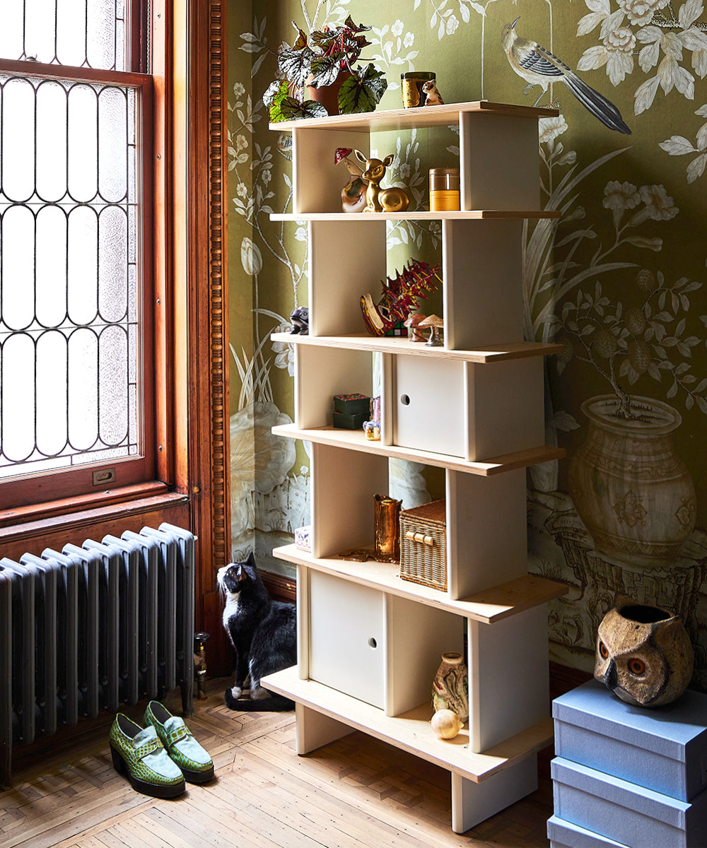 The Vertical Mini Library is adorned with decor, a cat, slippers, and boxes in a room featuring floral wallpaper and a stained glass window.