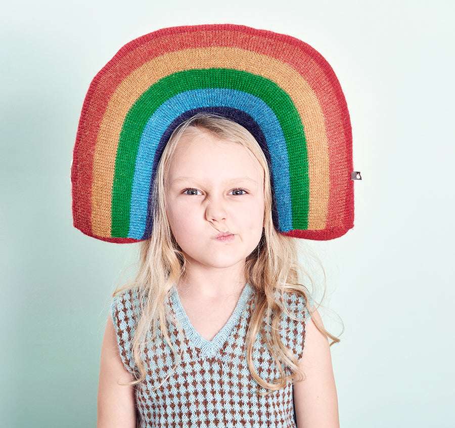 A child wearing a large, rainbow-shaped knitted hat makes a playful face. She has long blonde hair and is dressed in a patterned sleeveless top against a light blue background.