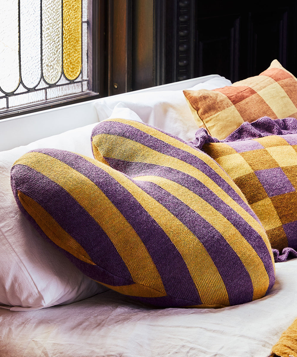 Striped Heart Pillow in yellow and purple on a bed with checkered pillows, stained glass window in the background.