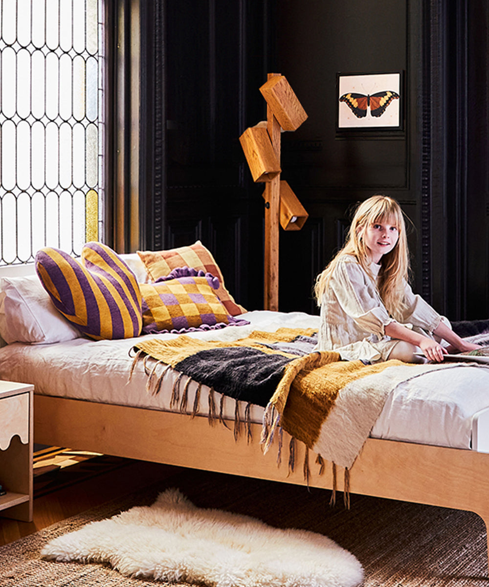 Girl sitting on a bed with Gingham Ruffle Pillows, colorful blankets in a cozy room with dark walls and a butterfly picture.