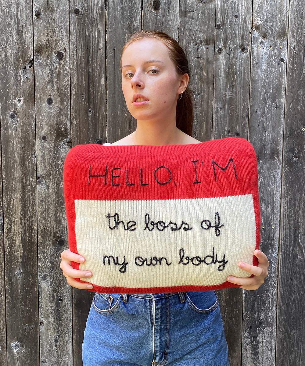 Person holding a Name Tag Pillow with "HELLO, I'M the loss of my own body" embroidered, standing against a wooden fence.