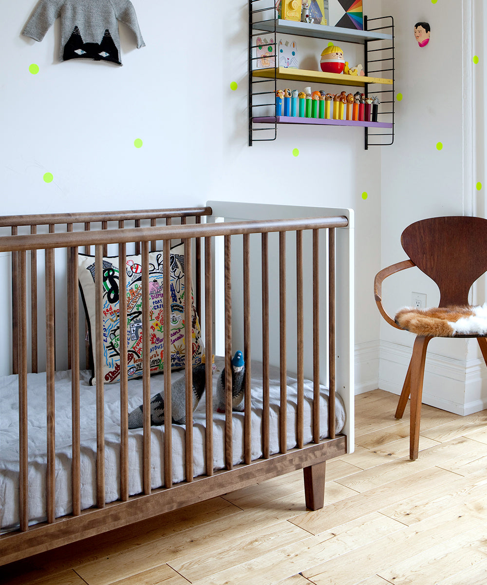 A cozy nursery featuring the Rhea Crib, colorful toys on wall shelves, and a wooden chair with a fur cushion on the floor.