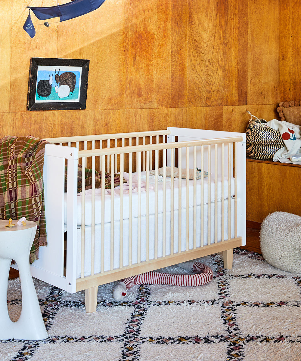 The Rhea Crib sits comfortably in a cozy nursery, surrounded by wooden walls, a plush toy, and a striped pillow on a patterned rug.