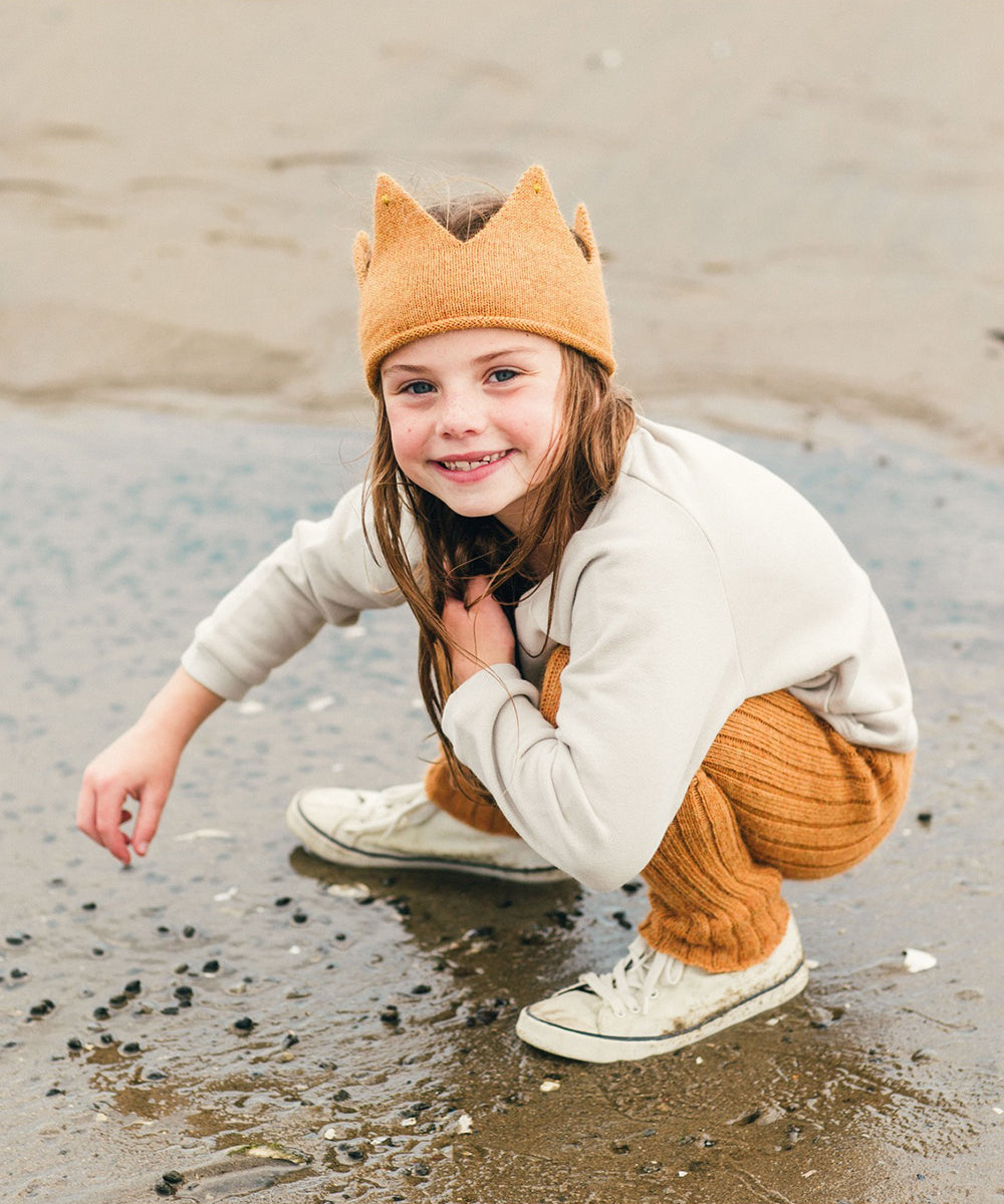 A child in a Gold Crown headband smiles while crouching on the sandy beach, dressed in a light sweater and orange pants.