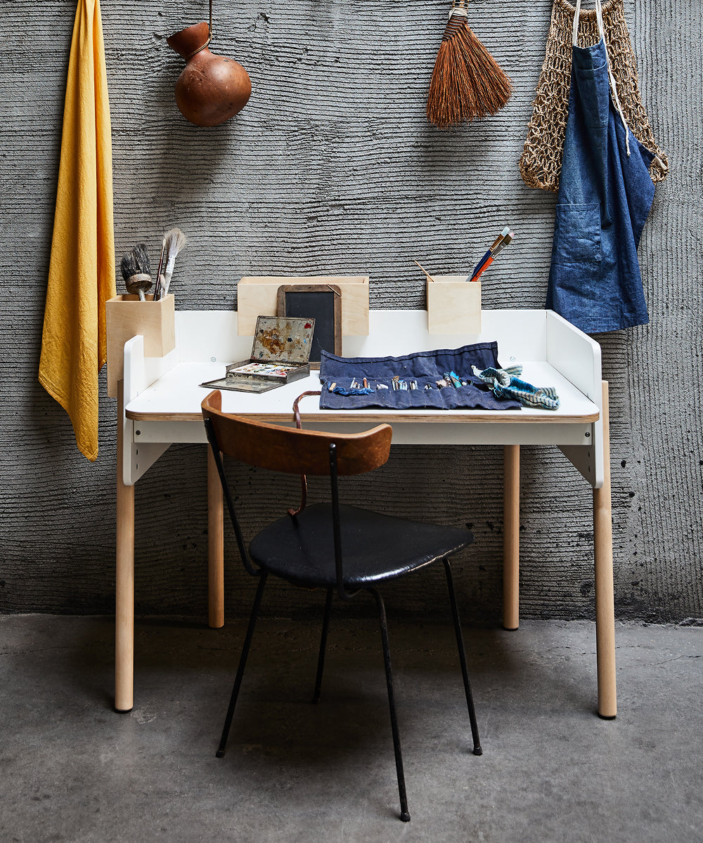 Cozy workspace featuring a Brooklyn Desk in white, complemented by a black chair and various tools; wall hangings include a broom and woven bag.