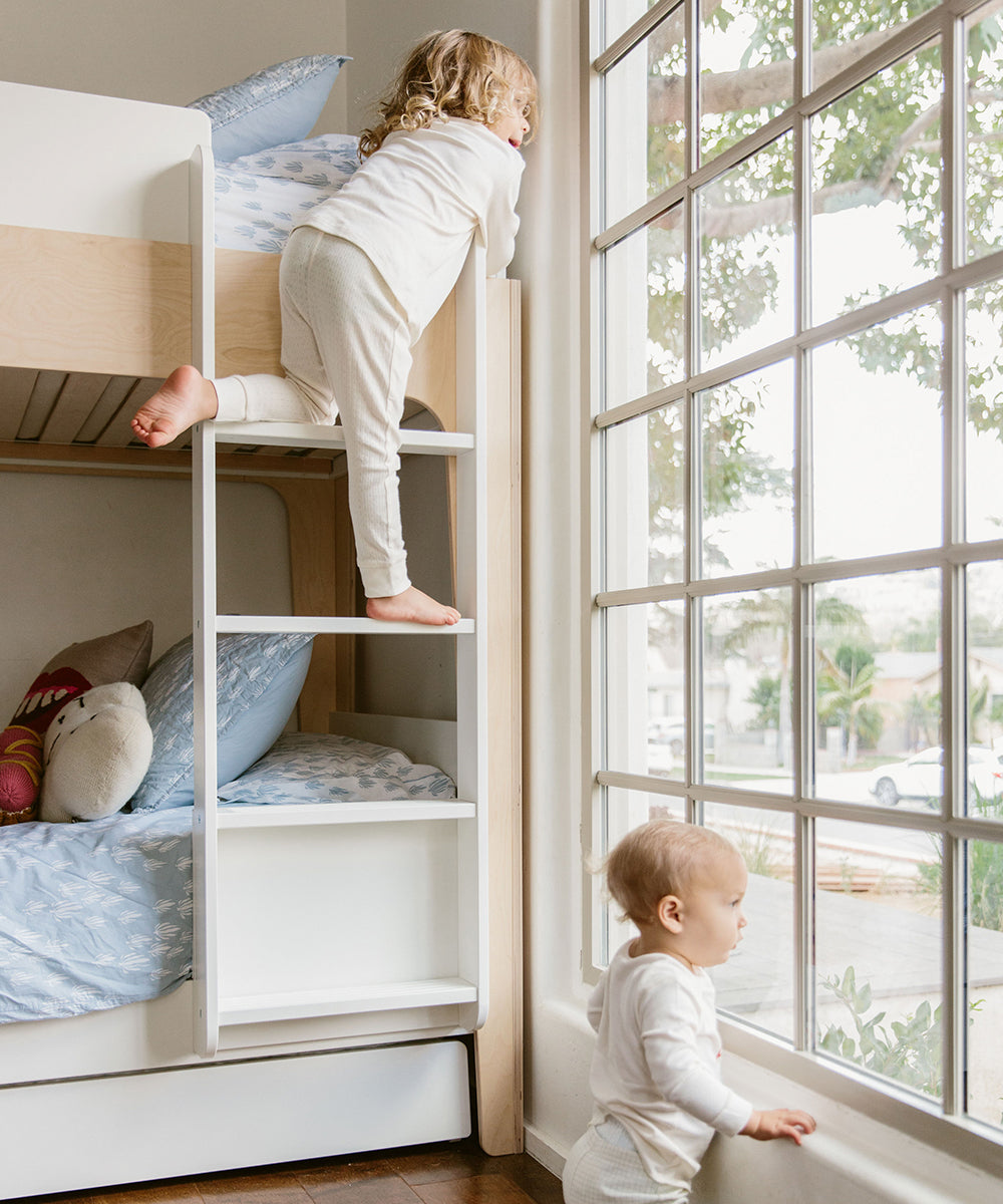 Two young children in pajamas are looking out a large window next to a Perch Trundle Bed & Ladder Conversion Kit inside a cozy room.