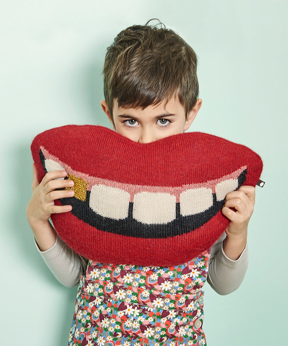 Child holding a "Mouth Pillow - Gold Tooth," a large pillow shaped like a smiling mouth, against a light green background.