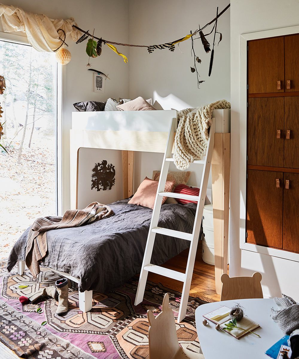 A cozy bedroom features the Perch Twin Bunk Bed, a vibrant rug, and leafy decor, all beautifully illuminated by light streaming through a large window.