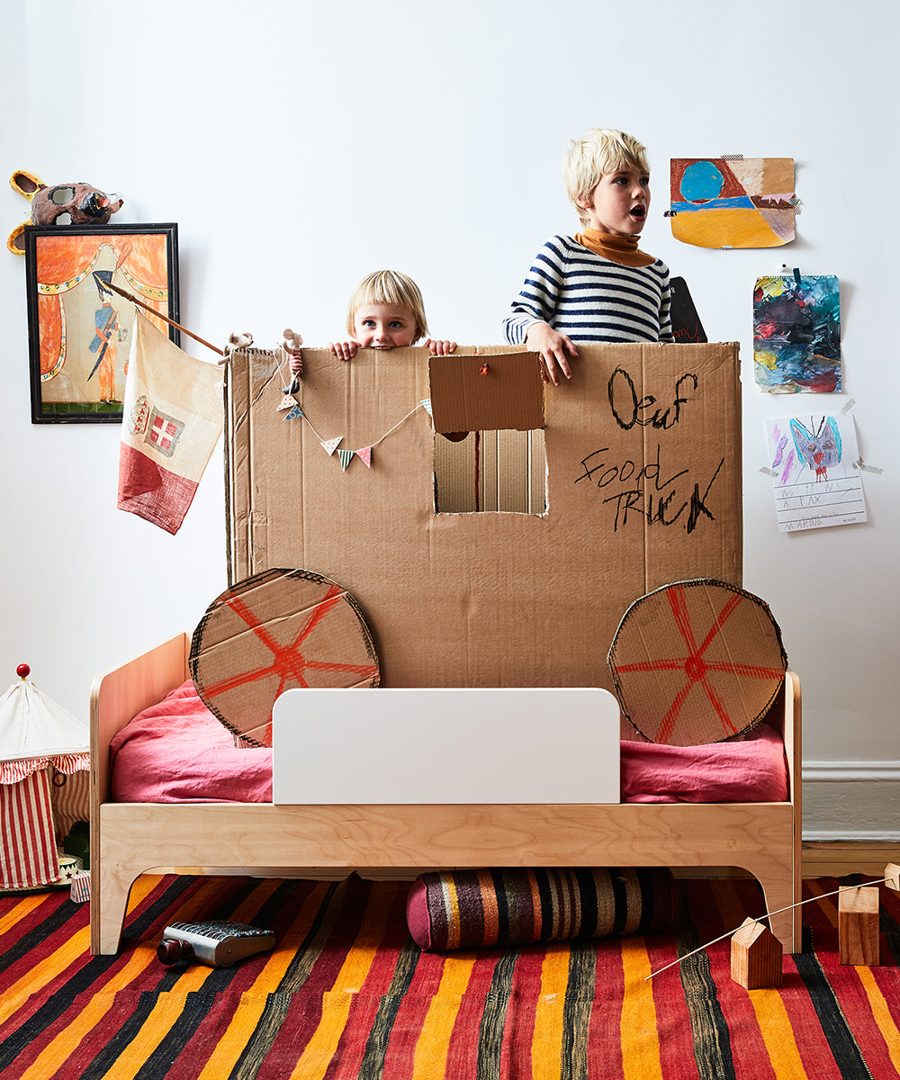 Two children play with a cardboard food truck in a vibrant bedroom featuring toys, drawings on the walls, and a Perch Toddler Bed.