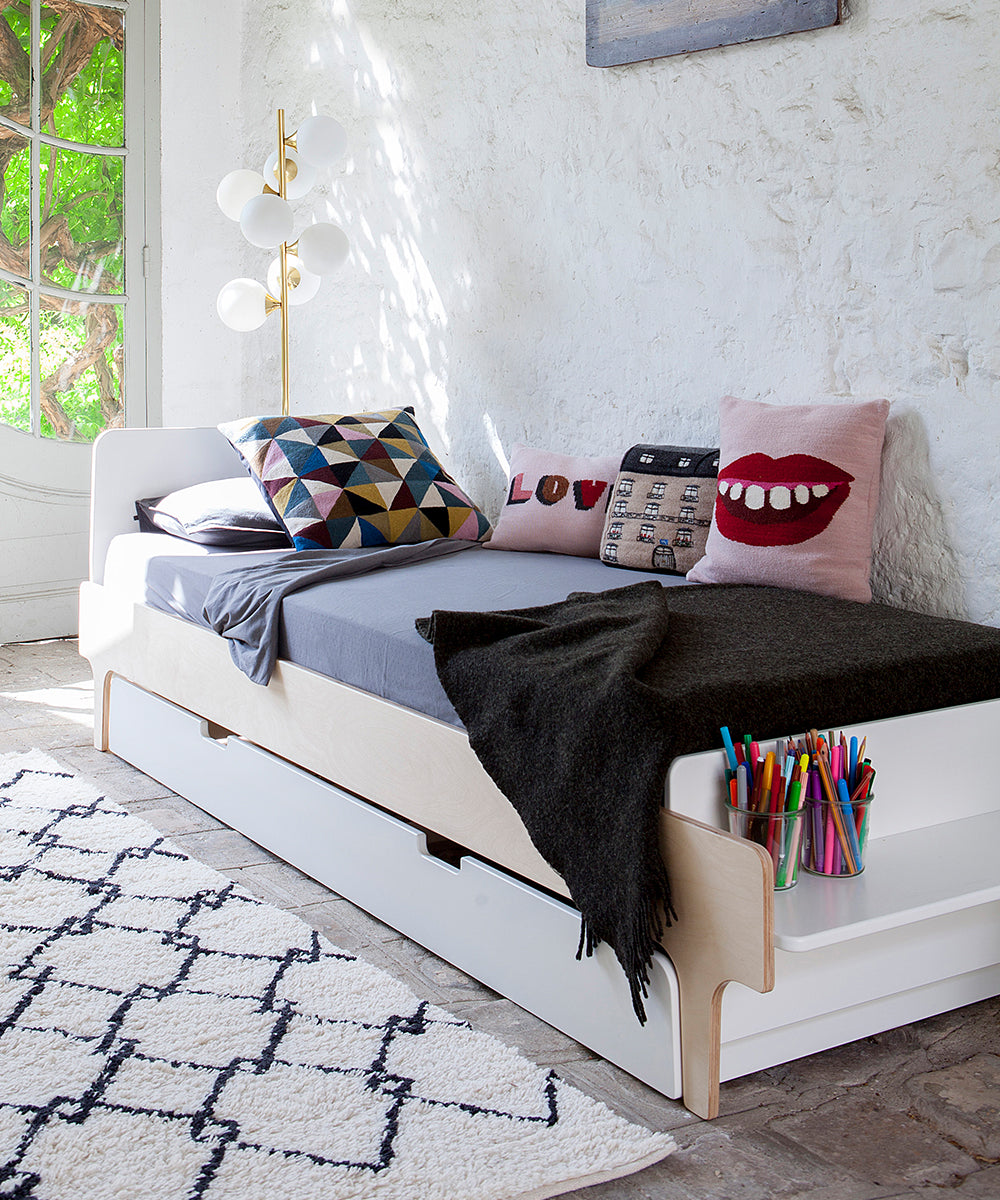 Room with a white River Trundle Bed, colorful pillows, a dark blanket, art supplies in a container, near a rustic window.