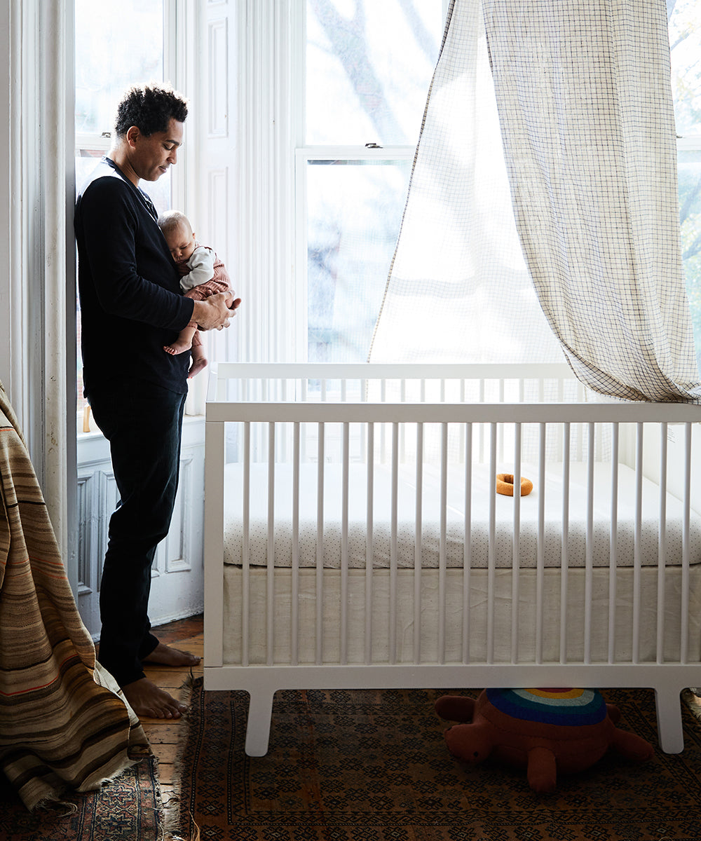 Someone holds a baby next to a sunlit Sparrow Crib made of birch hardwood, with a curtain partially covering the window.