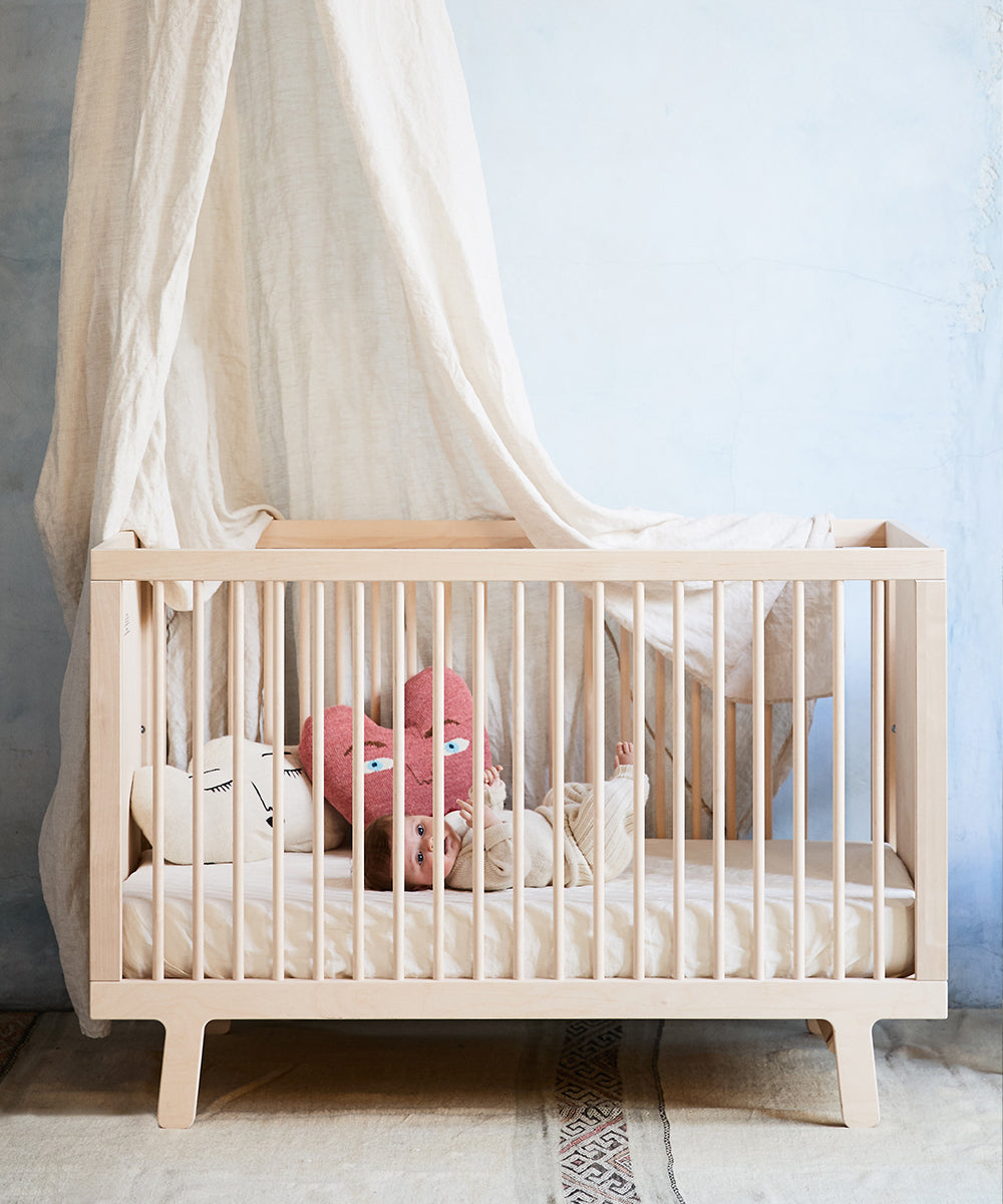 The Sparrow Crib, made from birch hardwood, holds a baby on a white mattress with plush, face-shaped cushions and a sheer canopy. This GREENGUARD Gold certified crib rests against a pastel wall.