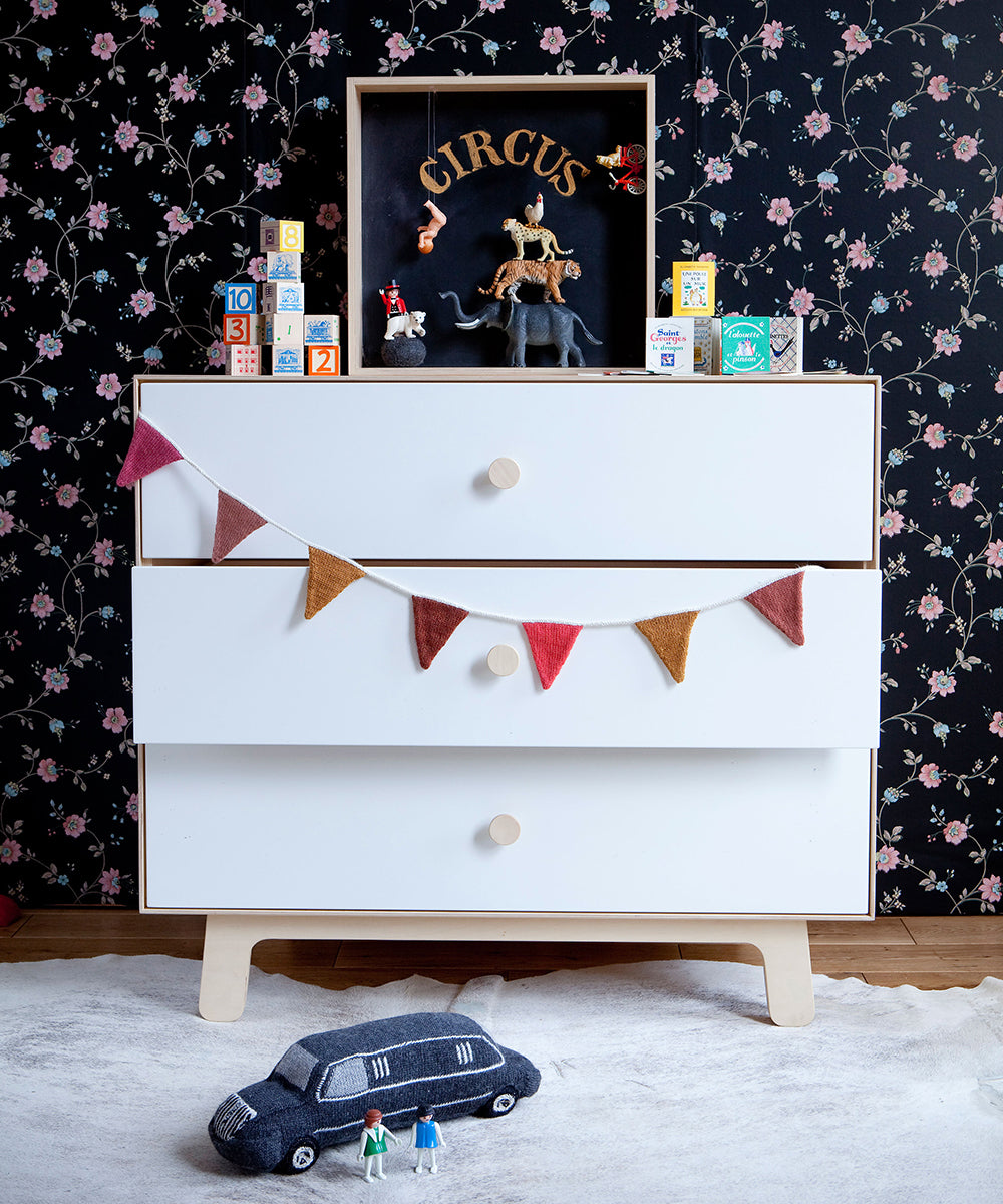 Sparrow 3-Drawer Dresser featuring circus-themed items, floral wallpaper backdrop, toy blocks on top, bunting across the drawers, and a toy car on the floor.