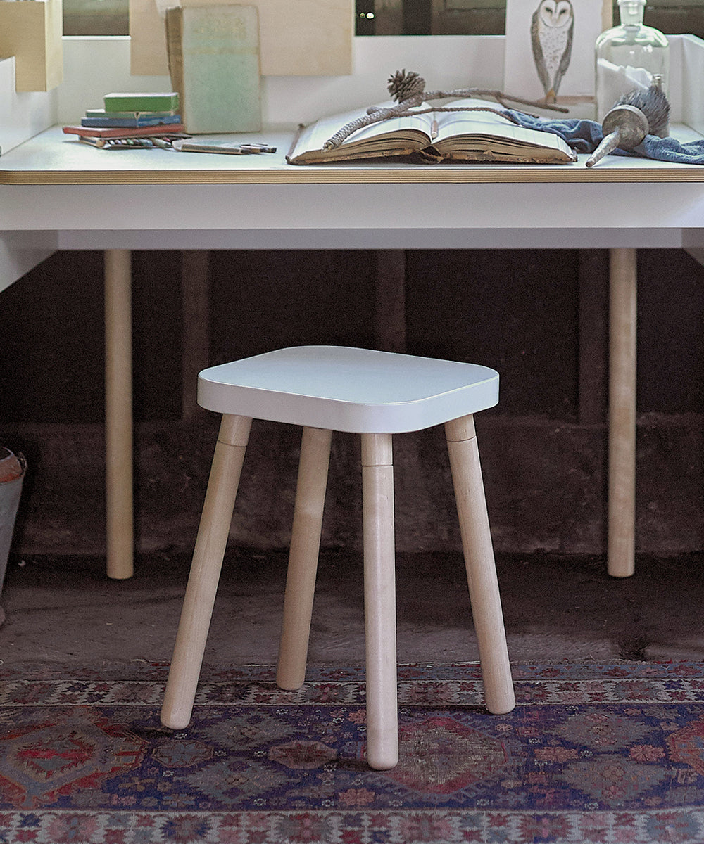 Square Stool with wooden legs in front of a desk, adorned with books and decor, sits on a patterned rug.