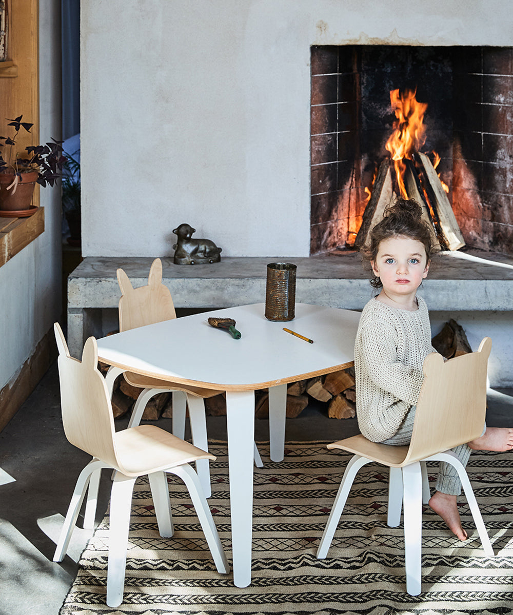 A child sits in a cozy, kid-sized space at the Play Table with bunny-shaped chairs, nestled before a lit fireplace on a rug crafted from sustainable materials.