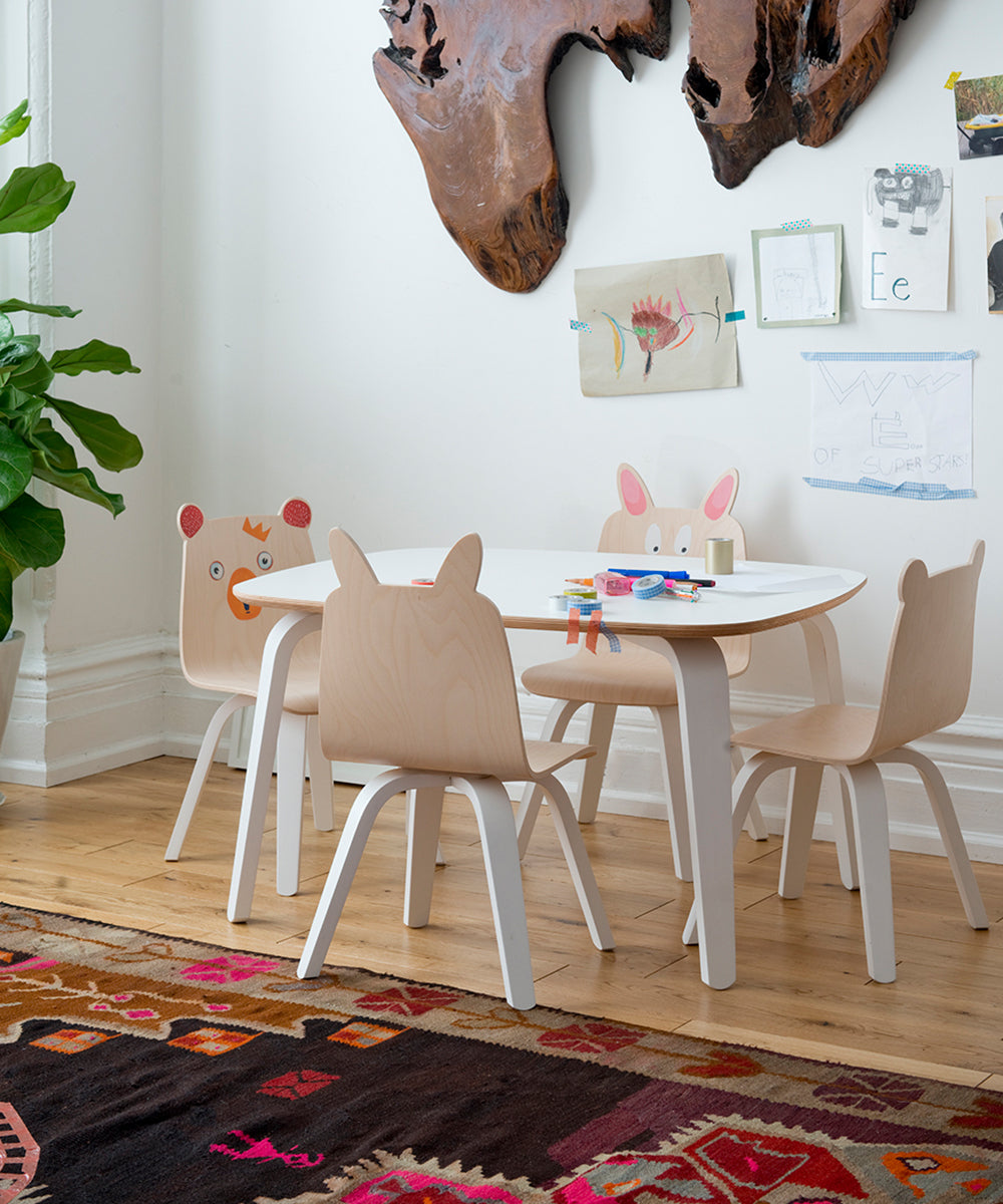 The kid-sized area includes a Play Table accompanied by chairs, some featuring animal designs. Art supplies and drawings are spread out, while a large leafy plant adds natures touch. The colorful rug enhances the wooden floor in this sustainable space.