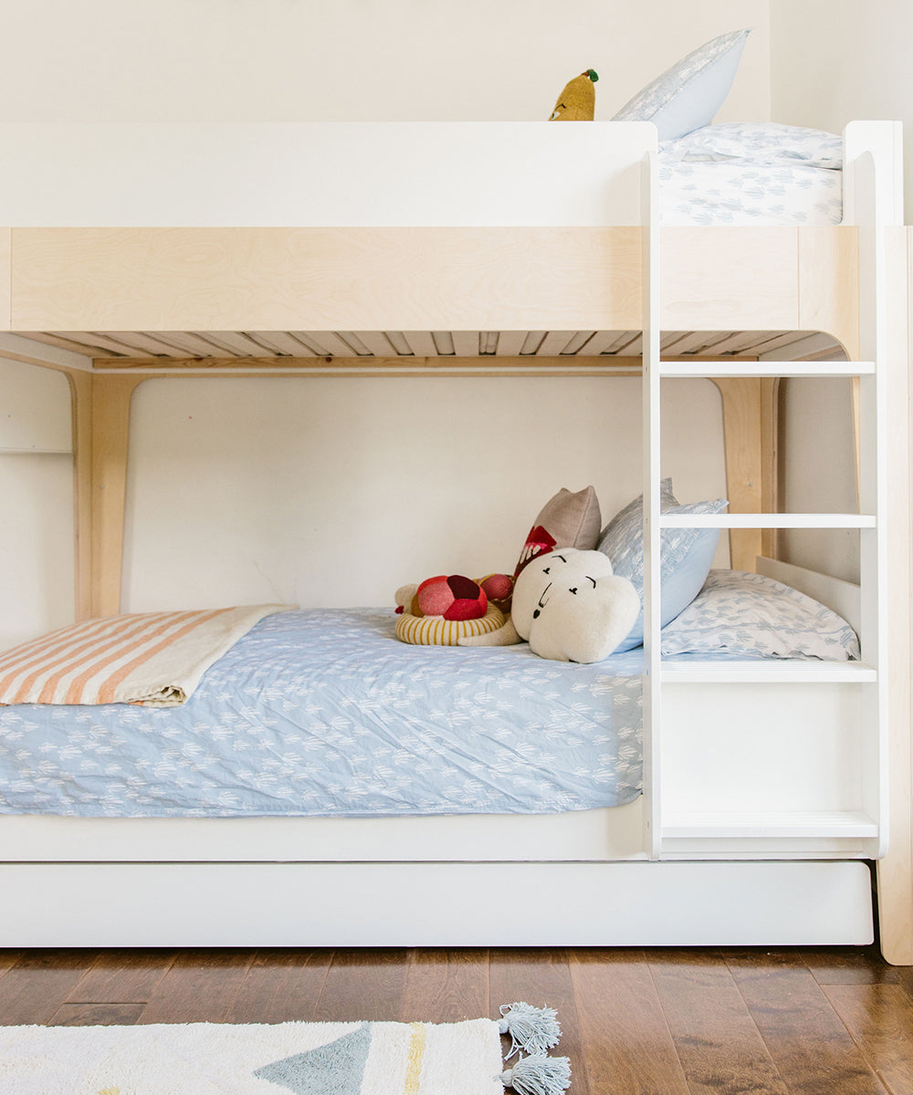 The Perch Trundle Bed & Ladder Conversion Kit features a wooden bunk bed with pastel bedding, stuffed toys on the lower bunk, and a white ladder on the right side.