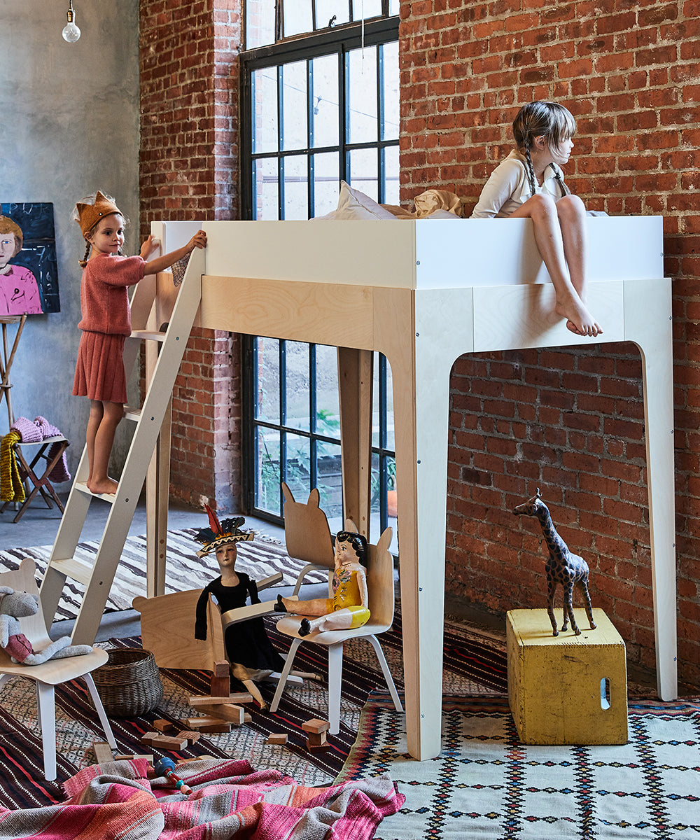 Two children enjoy their Perch Twin Loft Bed against brick walls; one sits on the bed while the other climbs the ladder. Toys and a giraffe figure are scattered across the floor beneath this versatile storage solution.