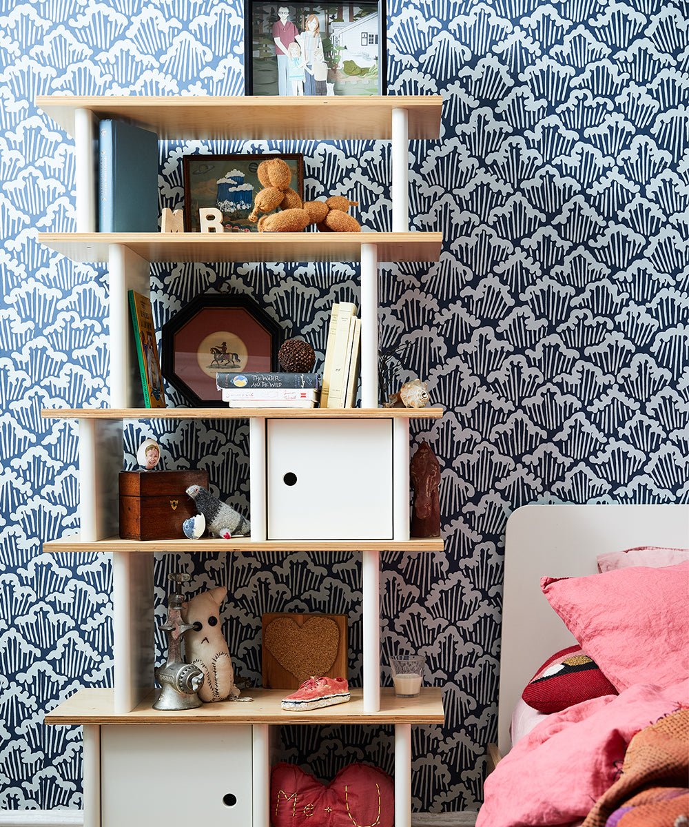 The Vertical Mini Library, adorned with books, stuffed toys, and decor, stands against a patterned blue wall with white storage boxes at the bottom.