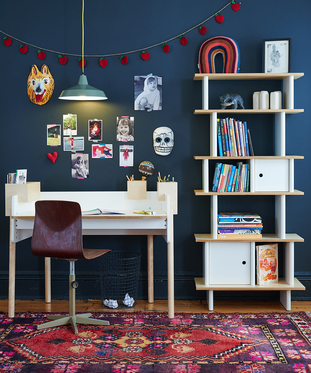 A colorful kids room featuring a Vertical Mini Library, desk, chair, books, wall art, patterned rug beneath a hanging light.